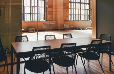 An empty meeting room with a long table and a row of chairs beside tall windows.